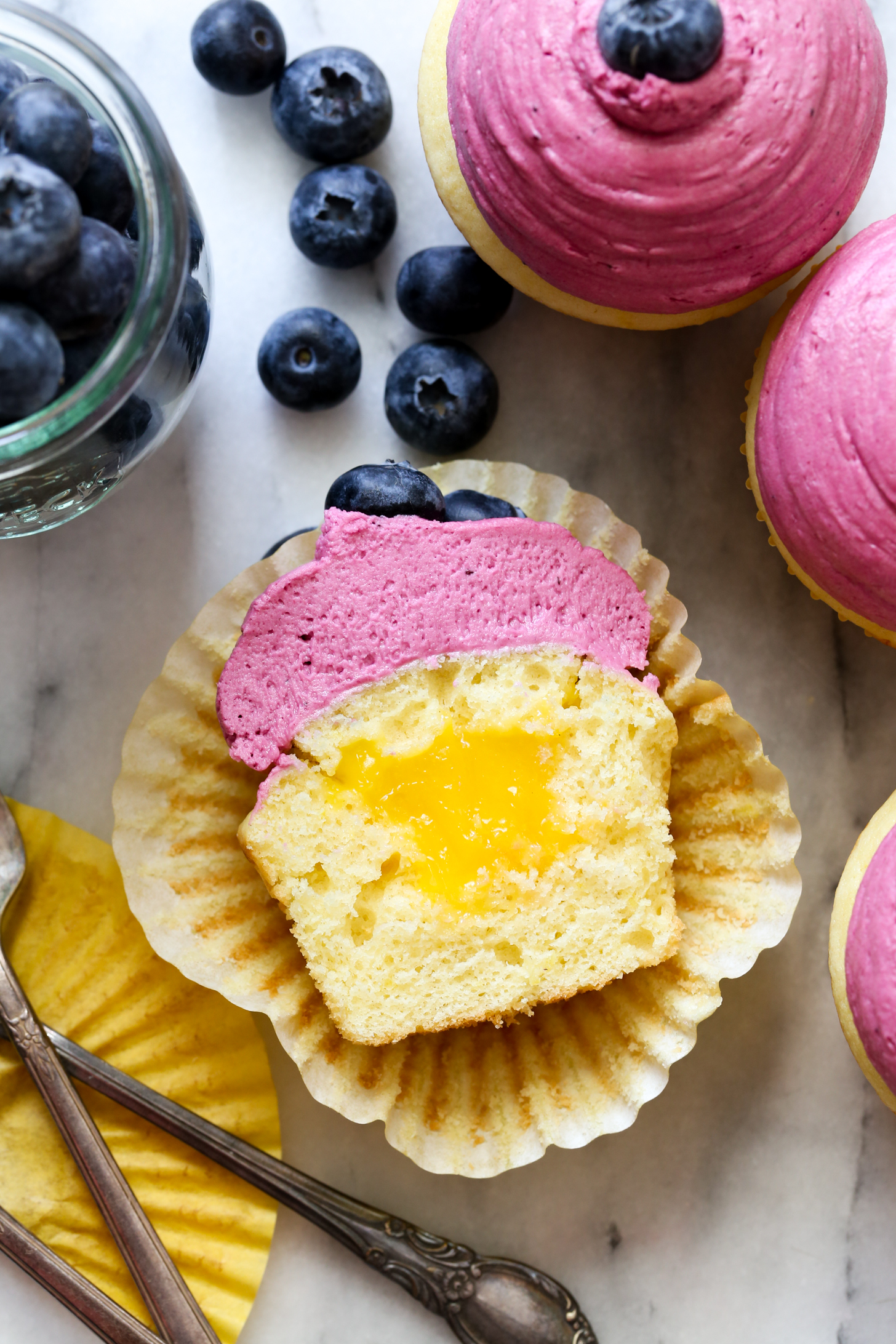 Moist and fluffy homemade lemon cupcakes with fresh lemon curd filling and blueberry buttercream frosting recipe from @bakedbyrachel A fun treat for any spring or summer occasion!